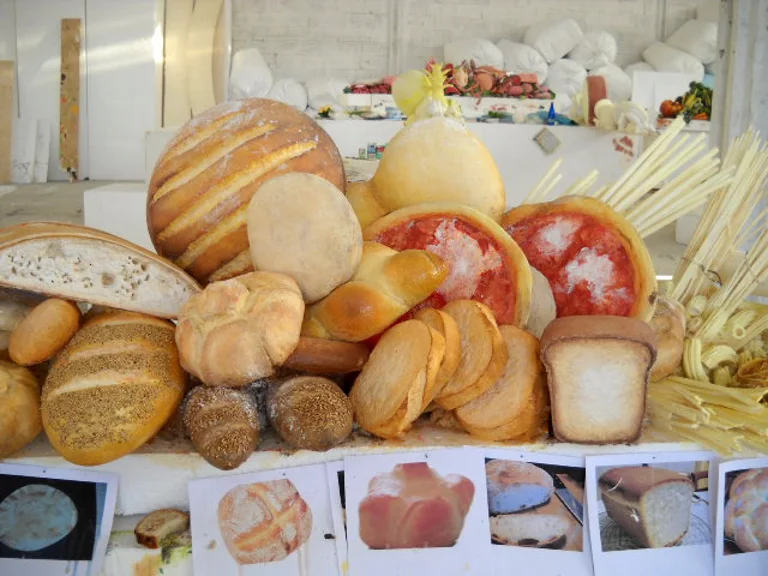 Still life of oversize sculpted bread for Occhio alla spesa: round loaf with crust striations, small michette rolls, braided brioche, tomato pizza, sliced baguette, sandwich bread loaf, donuts, focacce in the foreground; in the background a second counter with cured meats and cheeses