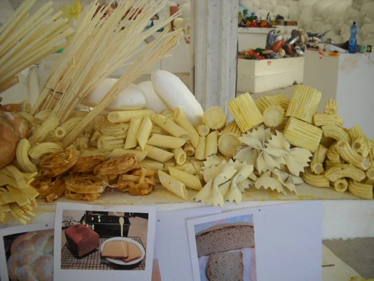 Sculpted pasta counter: bundles of tied yellow spaghetti, penne rigate, rigatoni, farfalle, brown rolled fettuccine, mafalde, on a white base; underneath, some reference photographs laid on the worktable
