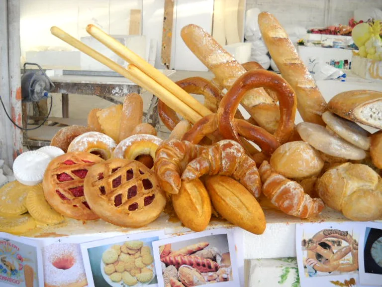 Second still life of bread and pastry: two upright baguettes, giant golden pretzels, red/brown lattice-topped jam tarts, croissants, focacce, donuts, sfogliatelle, two stand-up bread sticks like mechanic batons
