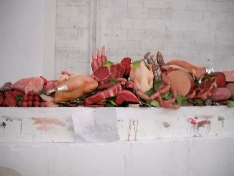 Hand-sculpted cured meats and meats counter: whole roast chicken, layered dry-cured hams, pink salami, pink frankfurters, mortadella, pink beef ribs with white fat, green salad leaves, on a white faux-brick stone base