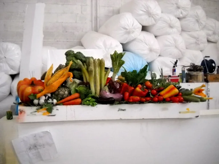 Counter of sculpted and painted vegetables: orange pumpkin, corn cobs with husk, bright orange carrots, dark green broccoli, celery, purple aubergines, red and yellow peppers, red cherry tomatoes, white and red onions, on a white base; bales of foam rubber in the background