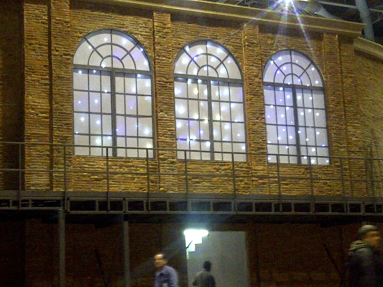 Side wall of the set with three large backlit arched windows: walls in sand-coloured faux stone ashlar, windows with round-arched stone frames, glass panes with a starlight effect of pale blue light points like stars in the night sky, central balustrade on the first floor