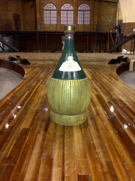 Detail of the set during finishing: close-up view of a black-and-gold twisted column with faux stone base, lattice wall with luminous circles in the background, gilded balustrade visible above, atmosphere of a TV studio with service lights