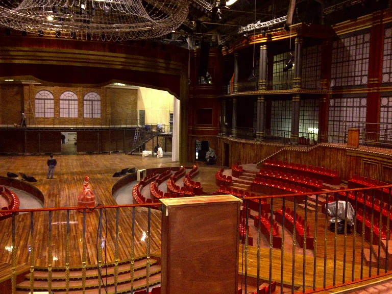 Interior view of the theatre set: central stage in light wood in the middle, double-tier loggia with box-balconies, black lattice structure on the walls, service lighting from the build, the back of the scene still being finished
