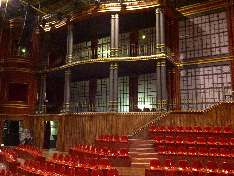 Frontal view of the theatre set rebuilt for a Giorgio Panariello show on Rai 1: three-tier boxes with central loggia, black-and-gold twisted columns alternating with lattice wall and luminous circles, gilded balustrades, backlit arched windows, in the foreground the stalls with padded red seats, central staircase to the stage