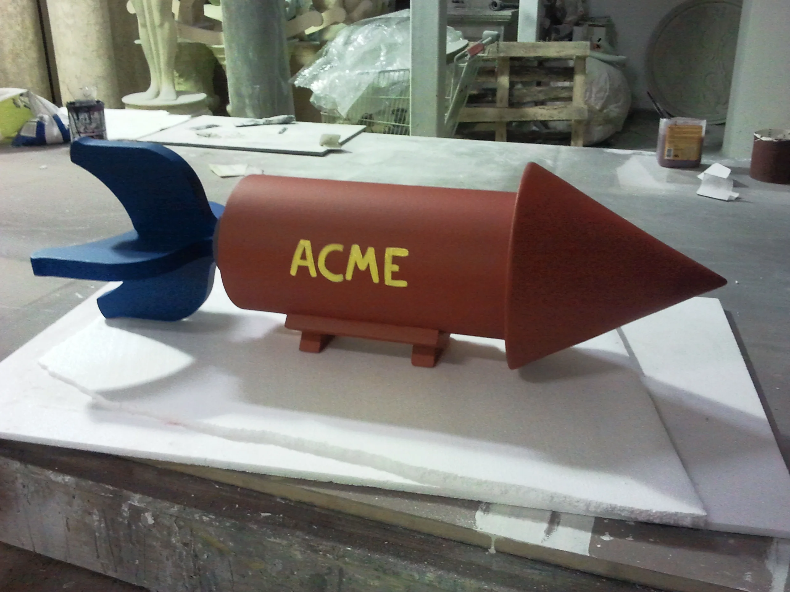 The ACME rocket bomb in the workshop during finishing: red cylinder still matte with freshly painted yellow ACME lettering, dark-red conical tip, blue fins, tripod legs; in the background the archive of sculptures in the Creation Art workshop