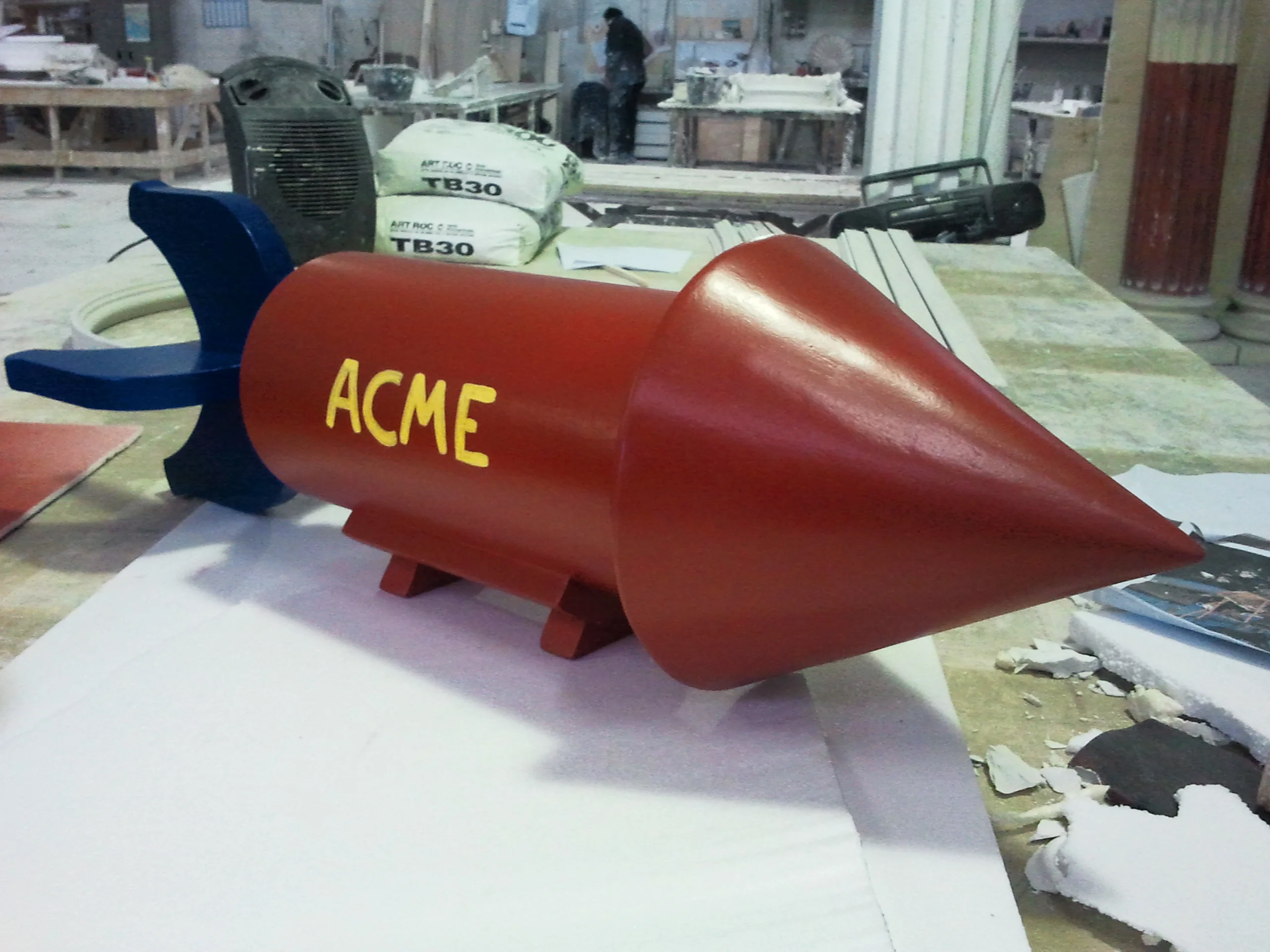 The ACME rocket bomb seen up close: bright red cylinder about a metre and a half long, 'ACME' lettering in block yellow on the belly, dark-red conical tip, blue stabiliser fins, tripod legs; in the background machinery and sacks from the workshop