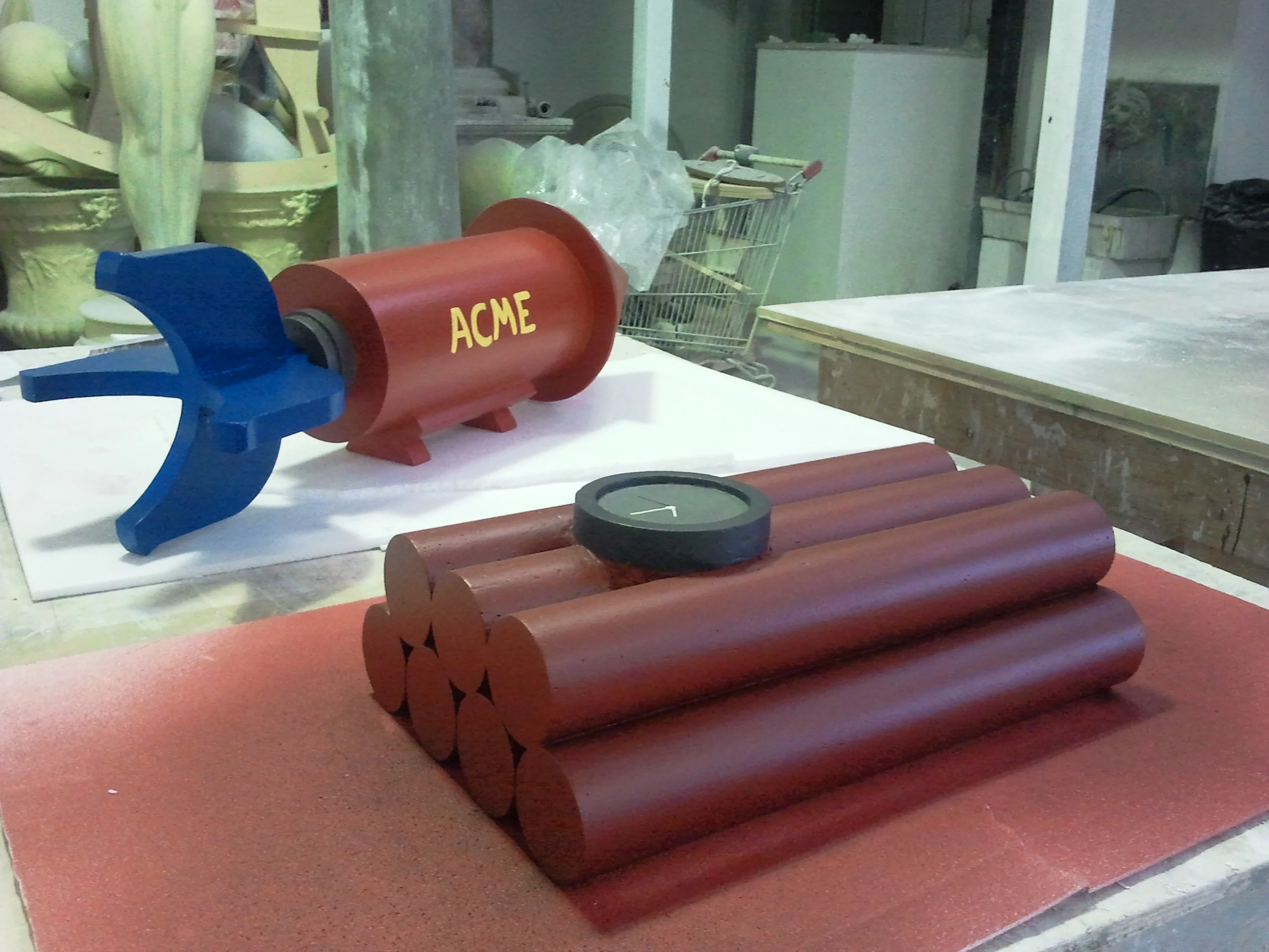 A pair of pieces on the workshop bench: top left the red ACME rocket bomb with blue fins, foreground right the dynamite with six red cylinders stacked in a bundle and round black detonator on top, resting on a red foam base