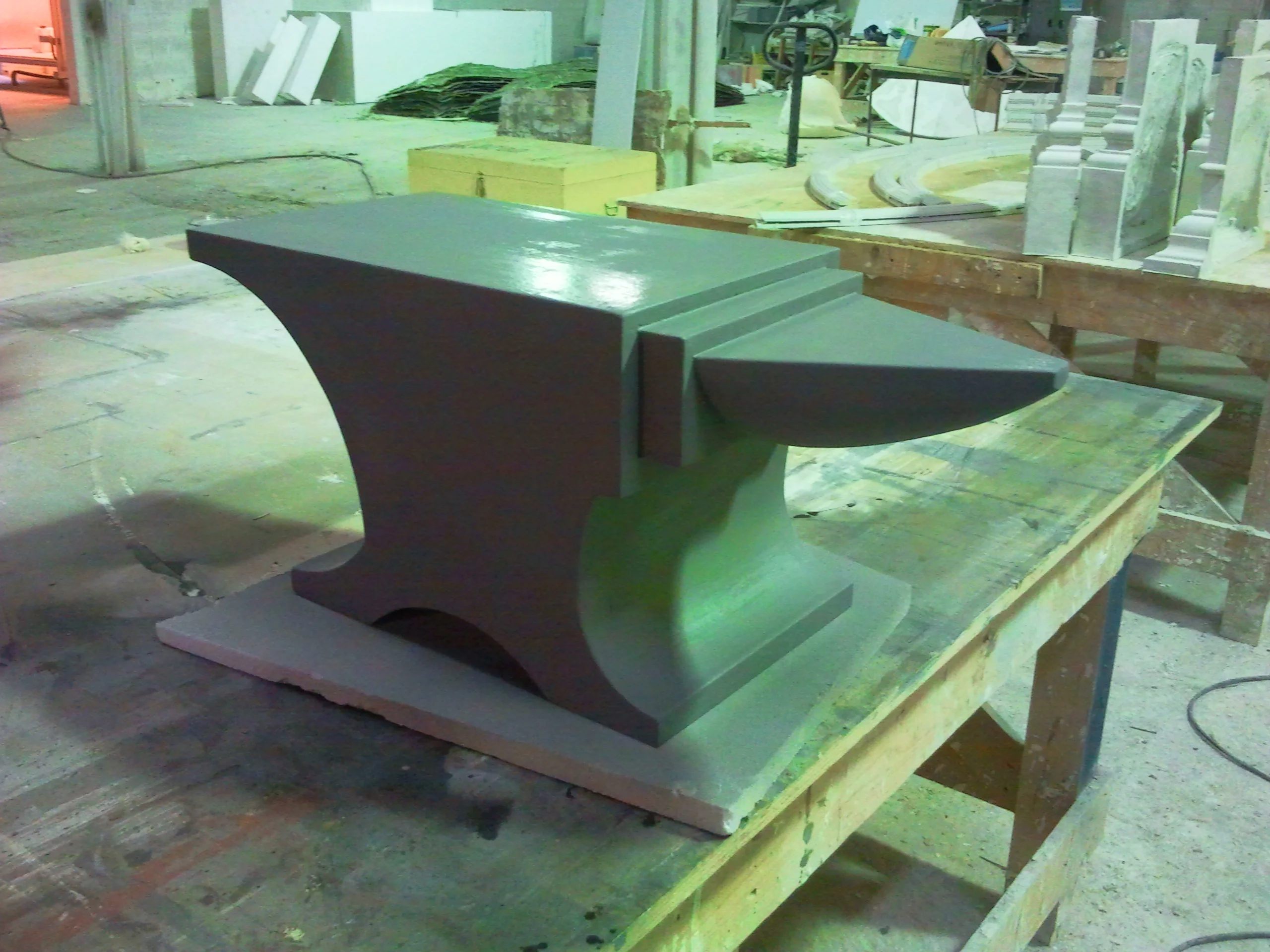 The ACME anvil sculpted in polystyrene and fibreglass, satin anthracite finish: classic blacksmith's anvil profile with flat upper face, side horn, recessed base; drying its primer in the workshop next to work tables