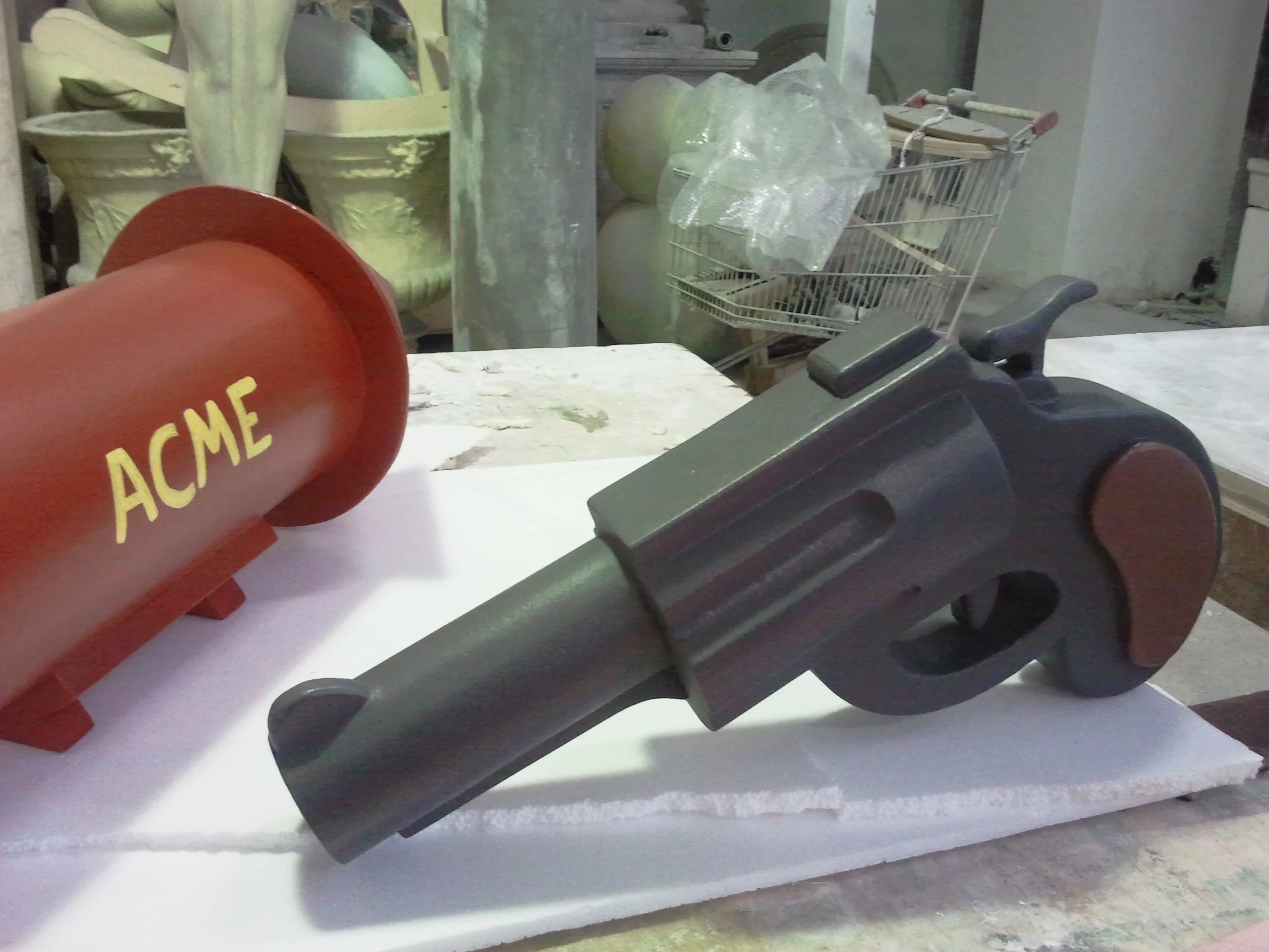 Steel-grey cartoon revolver resting next to the ACME bomb in the workshop: western revolver profile with cylindrical chamber, rounded grip, short barrel, satin grey finish; in the foreground a red ACME bomb on the left, in the background a trolley and sacks of material
