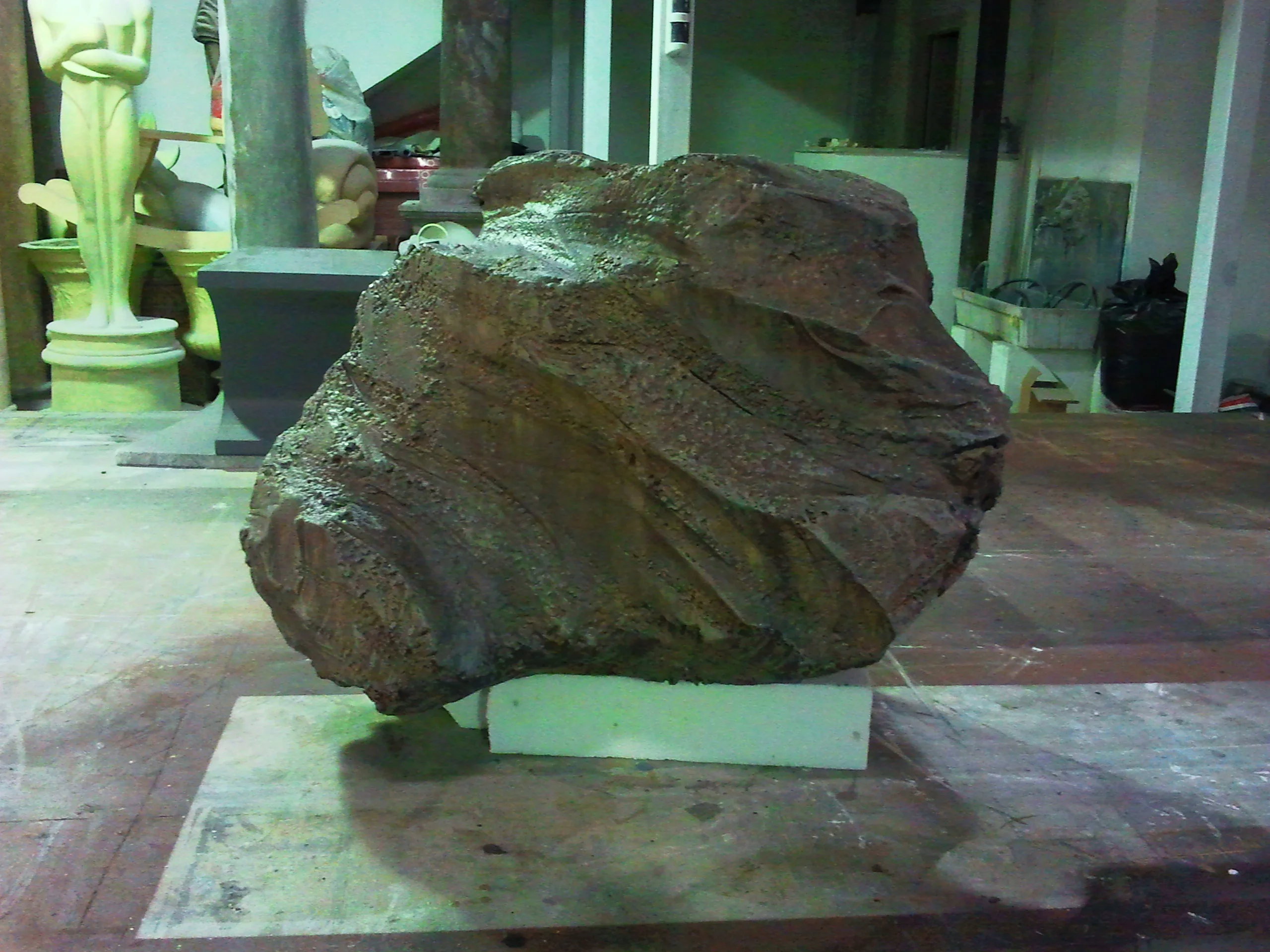 The ACME boulder sculpted in faux stone: irregular flattened-egg shape, eroded stone-effect surface in brown-grey, about a metre across, resting on a white polystyrene base in the workshop, in the background reproductions of white classical statues