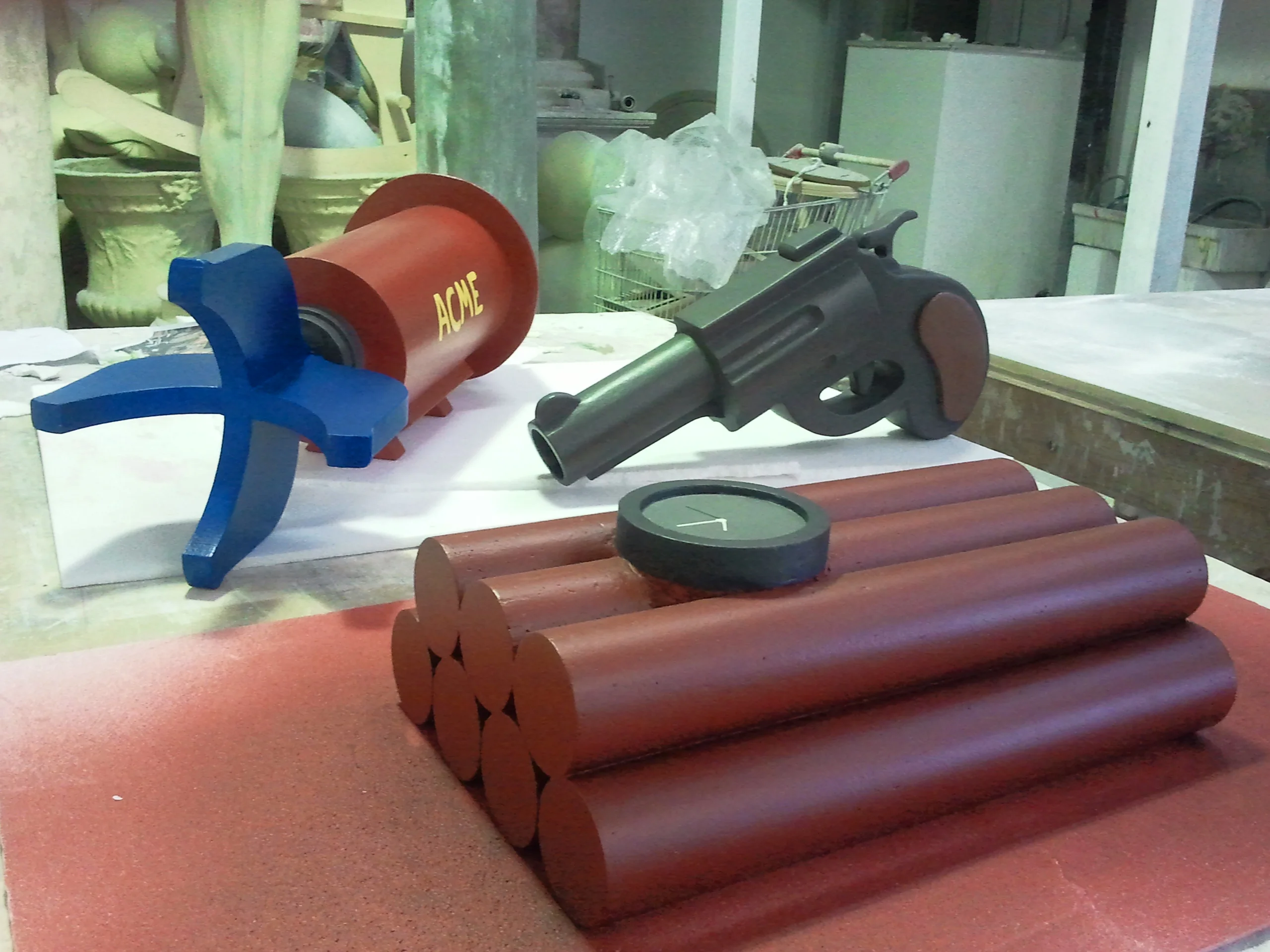 Trio of finished ACME props in the Creation Art workshop: on the left the red rocket bomb with yellow ACME lettering and blue fins, in the centre the grey cartoon revolver with cylinder and rounded grip, on the right the dynamite with six red cylinders and round black detonator on top; in the background drums and workshop materials