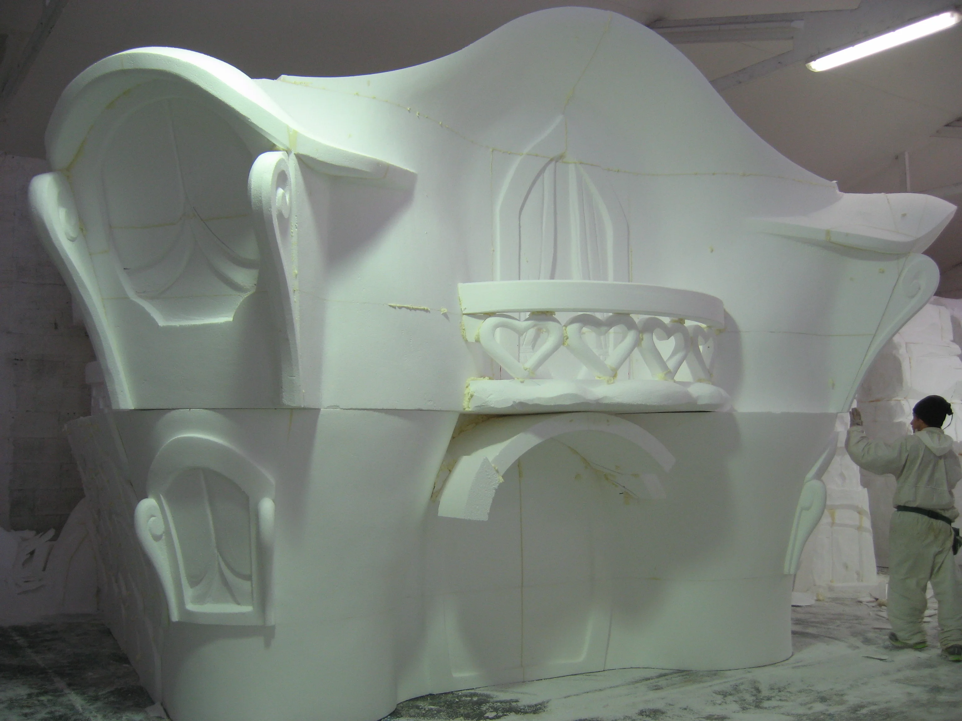 Detail of a Pixie House balcony sculpted in polystyrene: parapet with heart-shaped bas-reliefs, stylised Gothic-arched windows, wavy tiled roof, worker in white overalls on the right working in the background