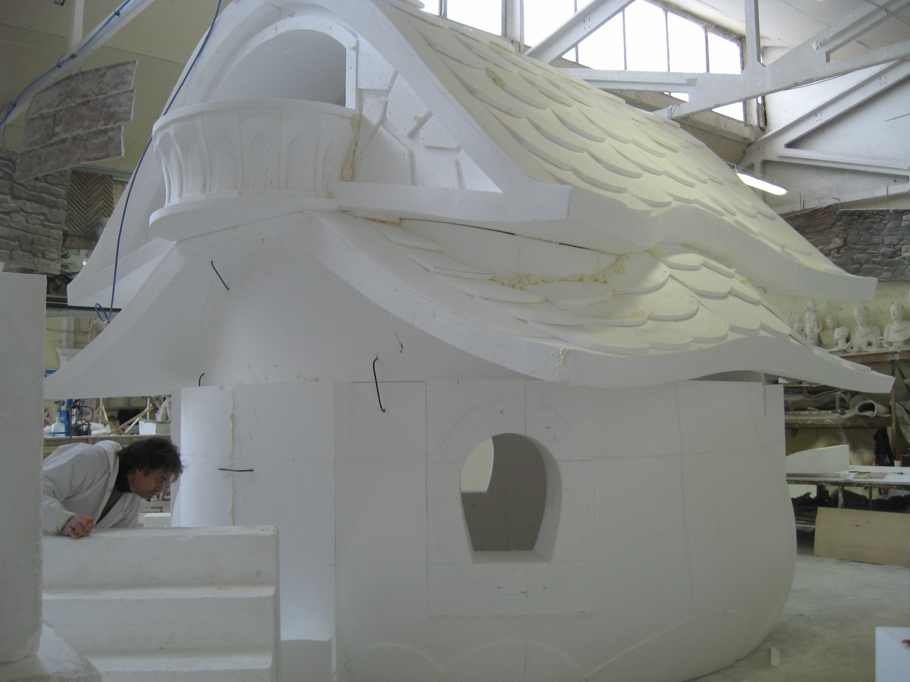 Pixie House under construction in the Creation Art workshops: sculpted in raw white polystyrene, still without finish, with a worker leaning against the side of the house working with tools; in the background industrial warehouse, polystyrene blocks, work in progress