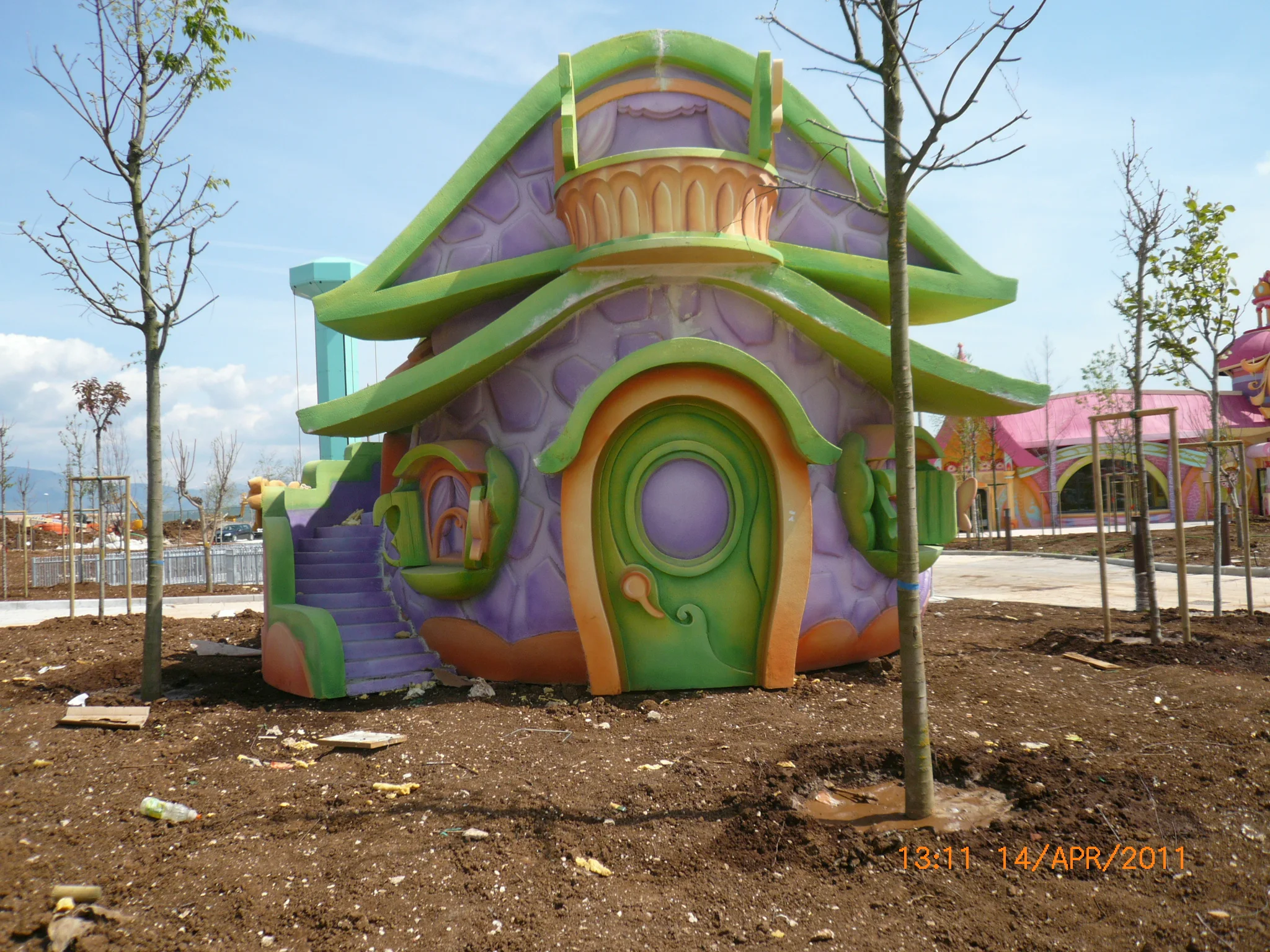 Another Pixie House installed in the park in April 2011: two-tone green and orange mushroom roof, purple walls decorated with geometric motifs, heart-relief balcony, drop-shaped window with curtain, orange slide coming out of the balcony; in the background a second pink and orange house, trees planted around