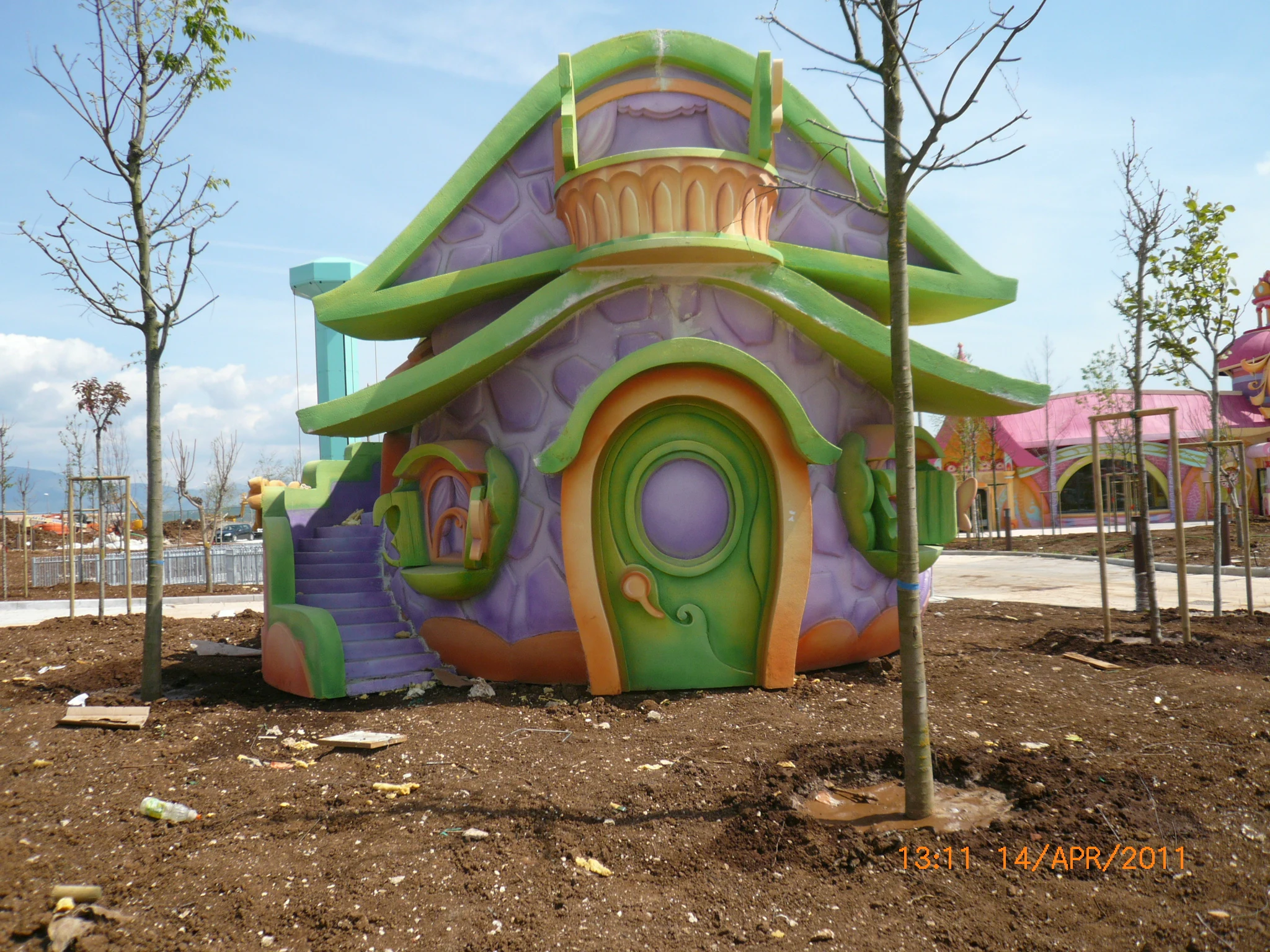 Pixie House installed in the park: green and orange mushroom roof, slide, balcony with hearts — saturated cartoon-colour finish
