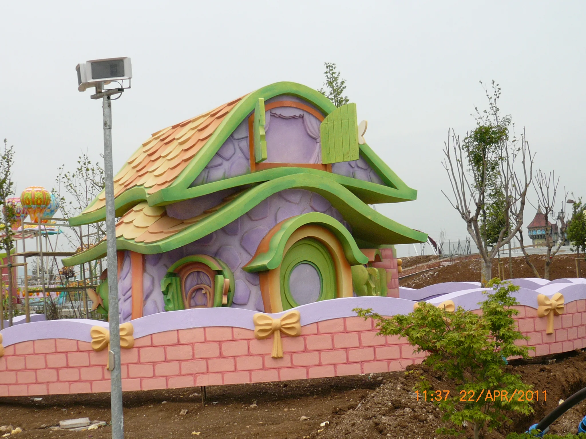 Overview of the completed Little Kingdom at Rainbow MagicLand: series of colourful Pixie Houses arranged around a square, newly planted trees, soft children's attractions, cartoon theme-park atmosphere