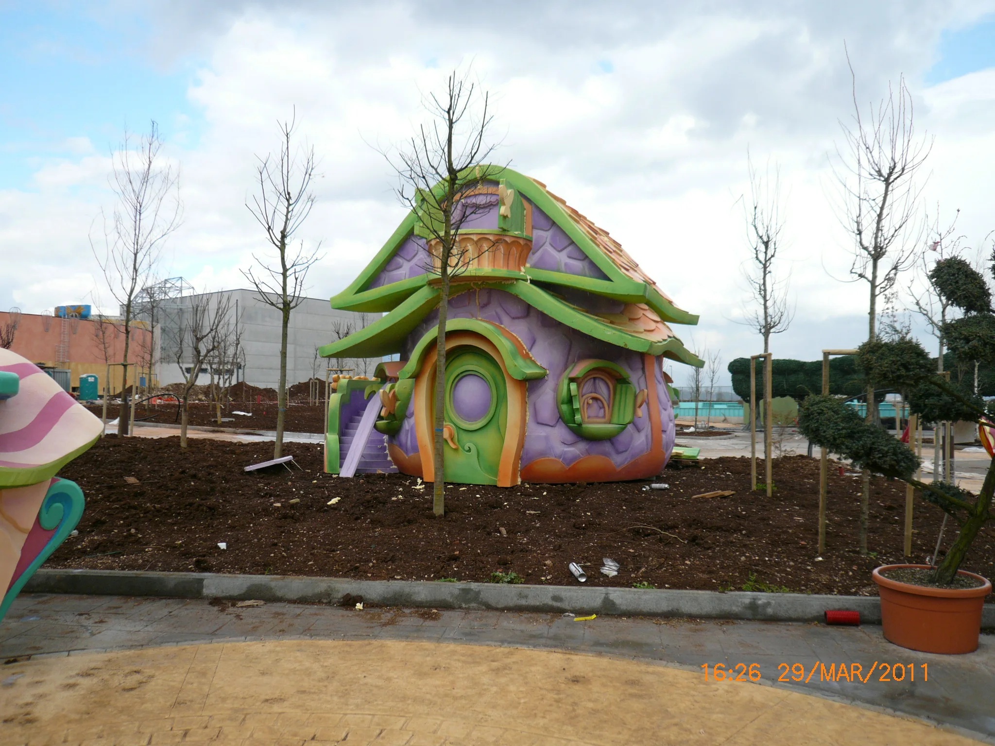 Pixie House installed at Rainbow MagicLand in March 2011: purple walls decorated with pink motifs, green mushroom roof with orange and yellow borders, oval windows with yellow frames, side orange slide; small trees newly planted around, pavement with construction material