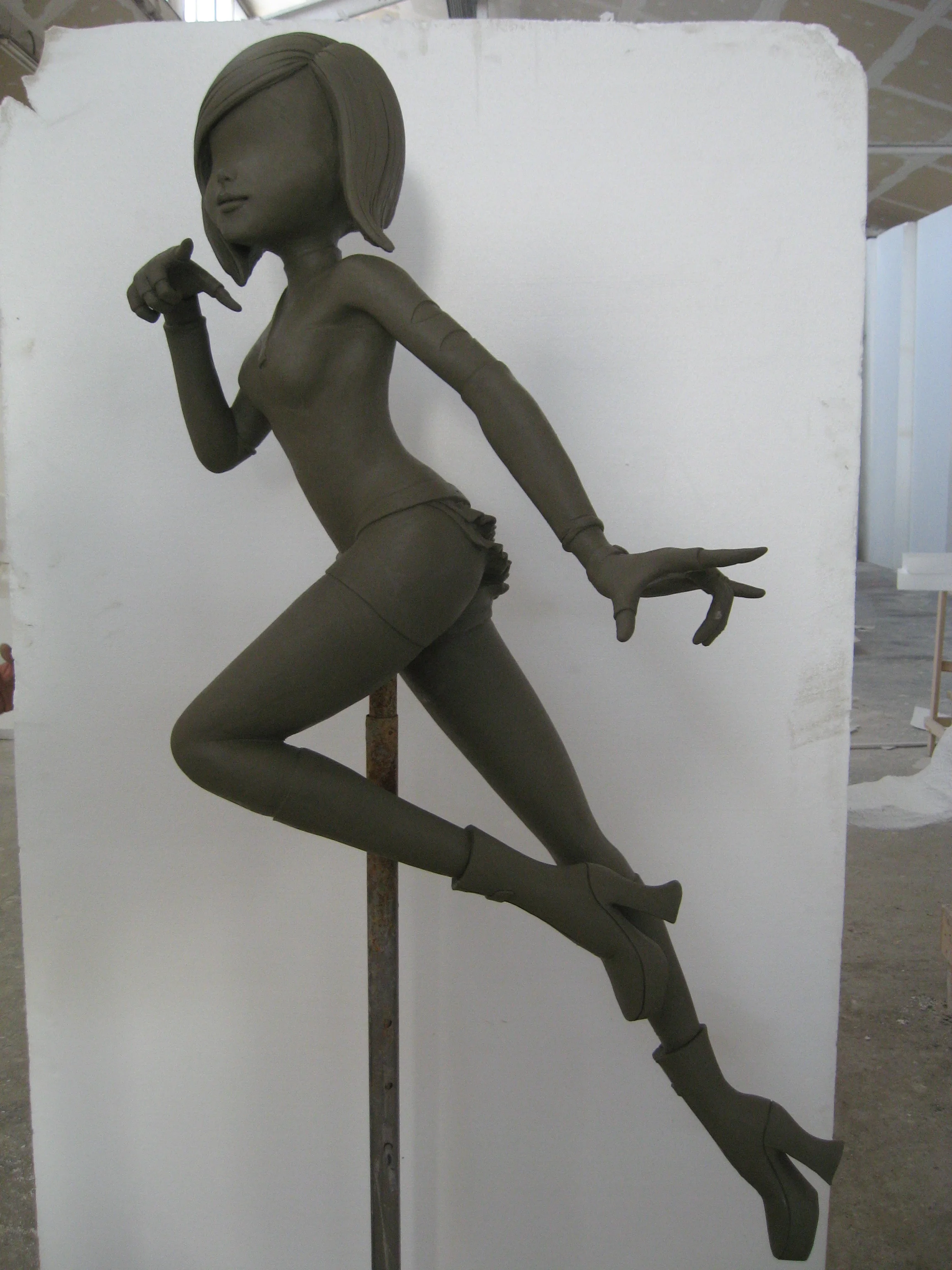 Statua di una Winx scolpita in argilla creta grezza nel laboratorio Creation Art: posa dinamica laterale con braccia alzate, gambe sollevate, capelli stilizzati a banda, montata su un palo metallico, pannello bianco di sfondo, capannone industriale visibile dietro