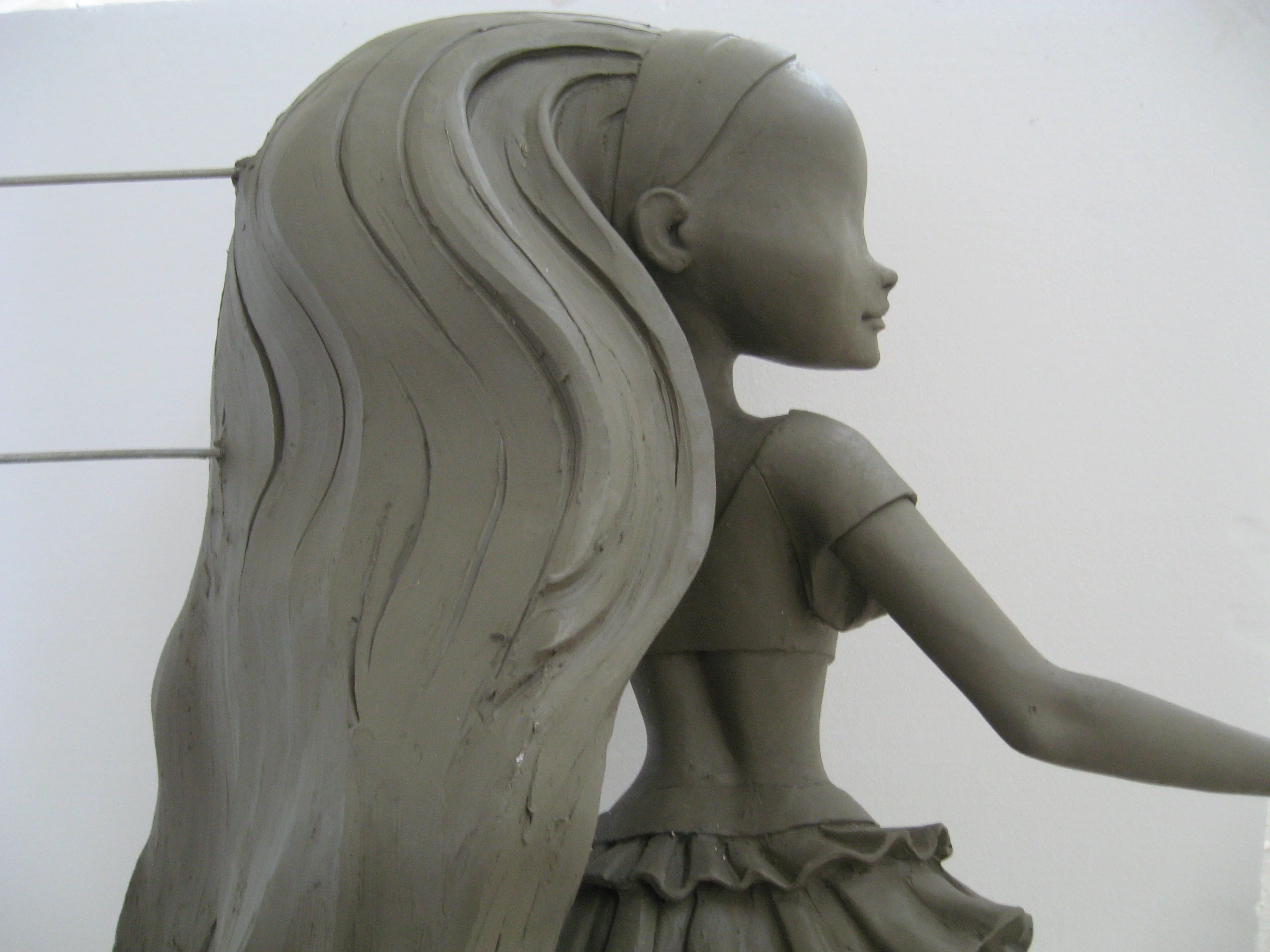 Profilo di una statua Winx in argilla: testa di tre quarti vista da dietro, capelli lunghi stilizzati a strati ondulati che le coprono la spalla, parete bianca del laboratorio con cavi tesi orizzontali