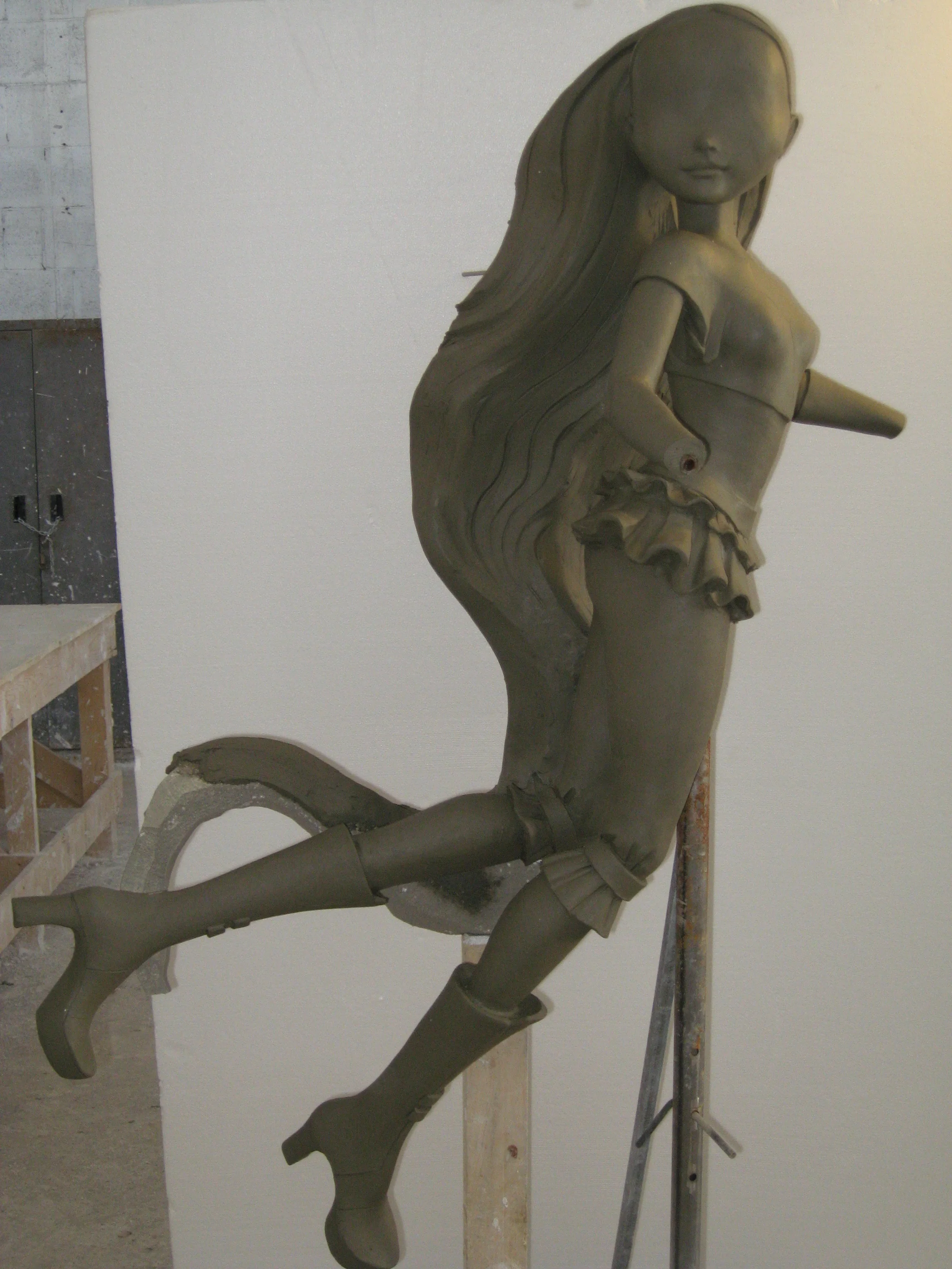 Statua intera di una Winx in argilla creta grezza, posa dinamica frontale con gambe sollevate come se fosse in volo, capelli lunghi mossi al vento, vestita con costume da fata e stivaletti, montata su palo metallico che regge il peso, parete bianca del laboratorio sullo sfondo