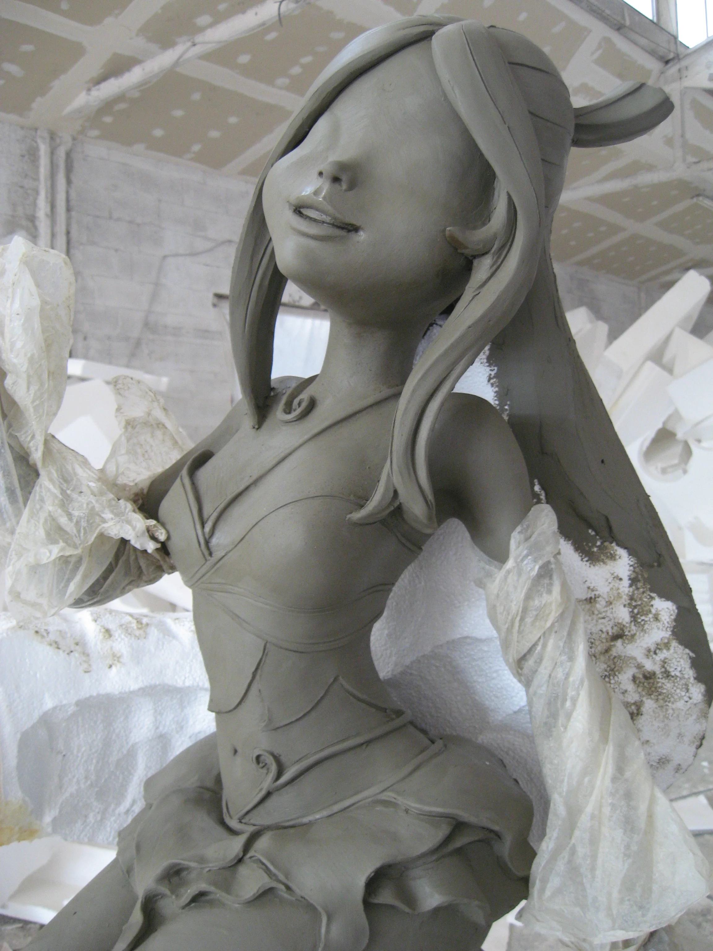 Primo piano della testa di una statua Winx in argilla creta grigia: viso simmetrico stilizzato, occhi chiusi, sorriso, capelli lunghi raccolti su un lato, fascia sui capelli; sullo sfondo pareti del laboratorio Creation Art e blocchi di polistirolo bianco