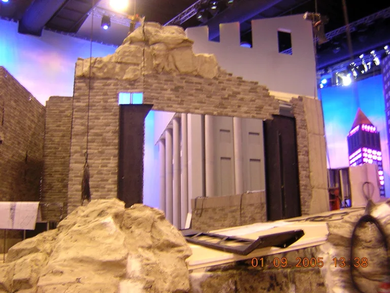 Construction of the urban ruin portal: grey brickwork sculpted in polystyrene + fibreglass, arches and jagged windows, at the top a crown of rocks, in the background the Art Déco skyscraper with magenta-lit spire, dated 1 September 2005
