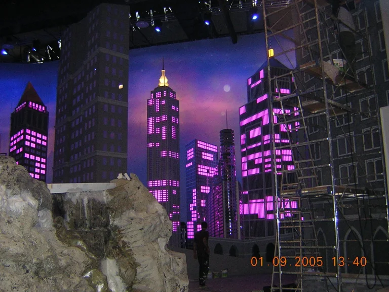Manhattan skyline rebuilt for RockPolitik in night light: sculpted skyscrapers several metres tall with magenta and violet back-lit windows, Art Déco building with central spire, white moon in deep blue sky, scaffolding on the right, rocky cliff in faux stone in the foreground, dated 1 September 2005