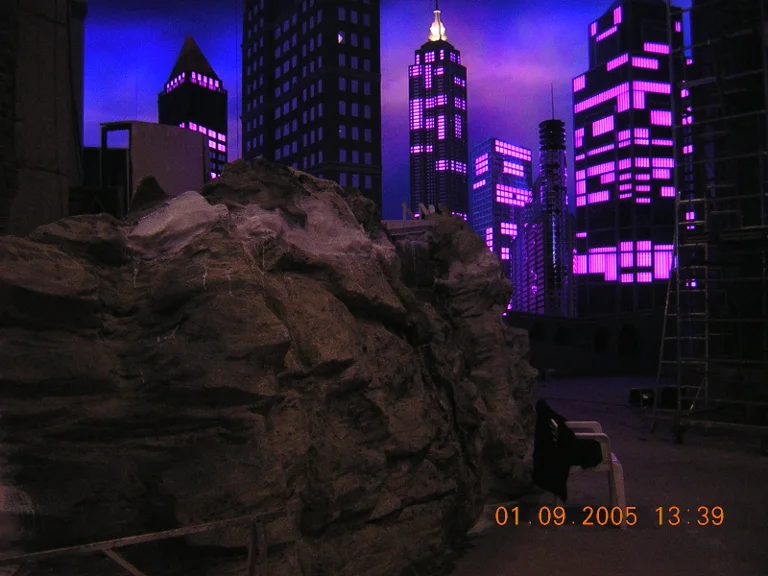 Side view of the night skyline of RockPolitik with a large sculpted rocky boulder in the foreground: faux stone rocks with white sculpted stalactites, skyscrapers in the distance with magenta-pink windows, Art Déco building with spire, ladder on the right of the build, dated 1 September 2005