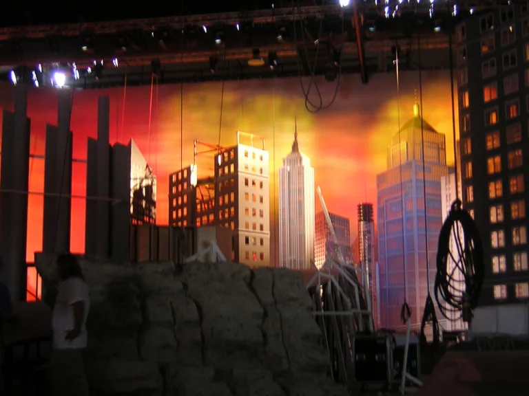The same skyline in warm daylight: orange-red sunset sky, stylised skyscrapers with the Empire State Building at the centre, in the foreground a grey faux stone rocky cliff, ladder standing on the left, scaffolding and overhead lighting trusses from above