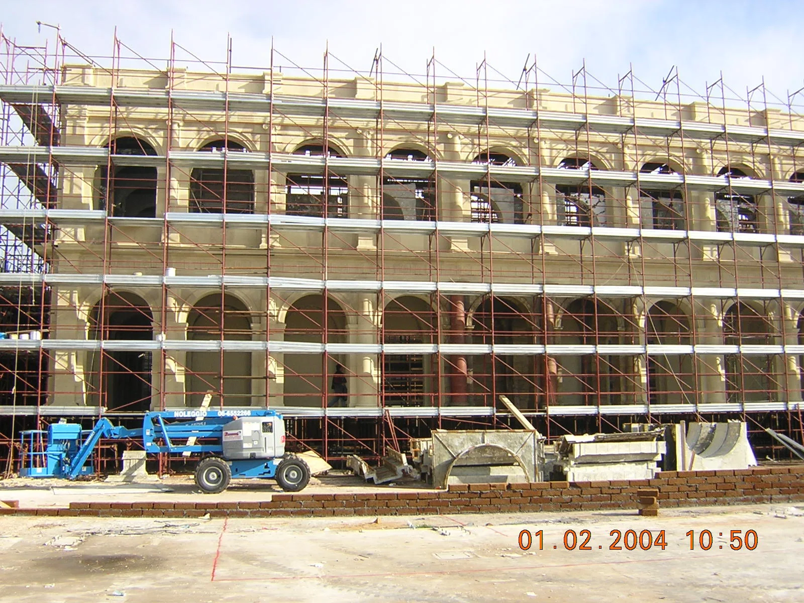 Set di Roma Antica a Cinecittà — cantiere del 1 febbraio 2004 per la serie ROME (HBO/BBC): facciata monumentale a due piani con ponteggi metallici che la avvolgono completamente, archi sul piano superiore visibili attraverso i ponteggi, una gru ad accesso a piattaforma blu sulla sinistra, mattoni e pannelli posati sul piazzale