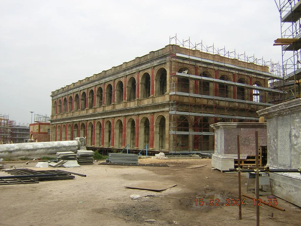 Set di Roma Antica — cantiere del 15 febbraio 2004: edificio monumentale a due piani d'archi ormai con muratura grezza e prima posa delle colonne in marmo finto rosso, ponteggi sulla destra ancora in posto, sulla sinistra una grande colonna marmorea scolpita appoggiata a terra, sul piazzale frammenti di pavimentazione, terra battuta polverosa, cielo grigio invernale