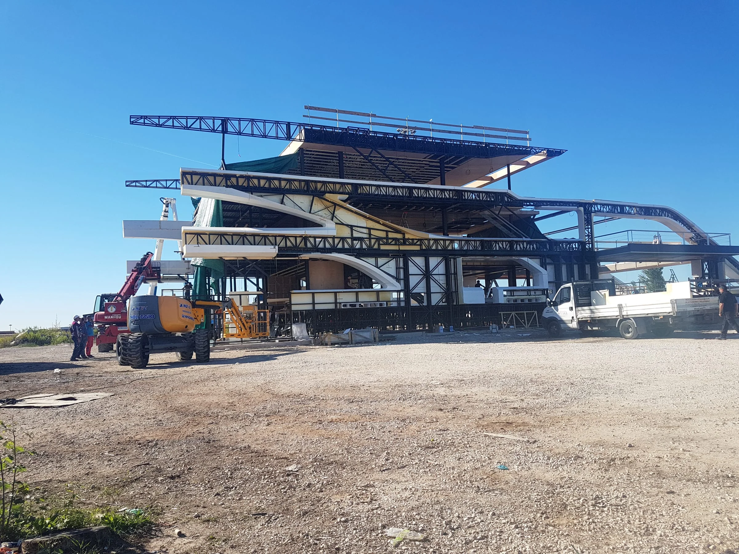 Mega-yacht scenografico per 6 Underground in cantiere il 27 settembre 2018: imponente impalcatura nera in metallo a forma di yacht, pannelli bianchi già installati ai primi due ponti, gru telescopica gialla con braccio rosso al lavoro sopra, escavatore bianco e mezzo a destra, terreno sabbioso del piazzale, cielo azzurro