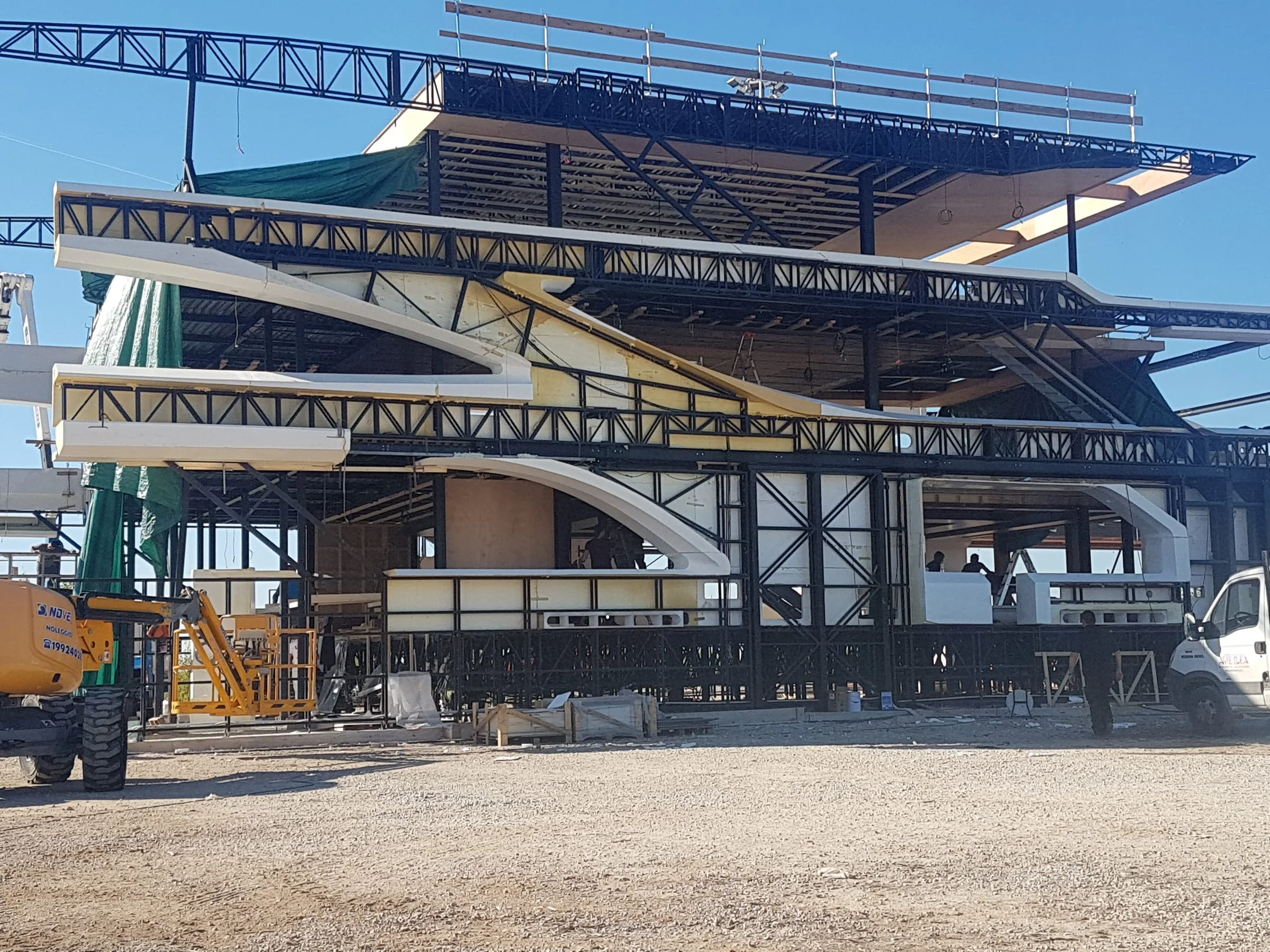Altra vista del mega-yacht scenografico in cantiere: dettagli dei pannelli bianchi installati sui ponti, struttura metallica visibile, mezzi e operai