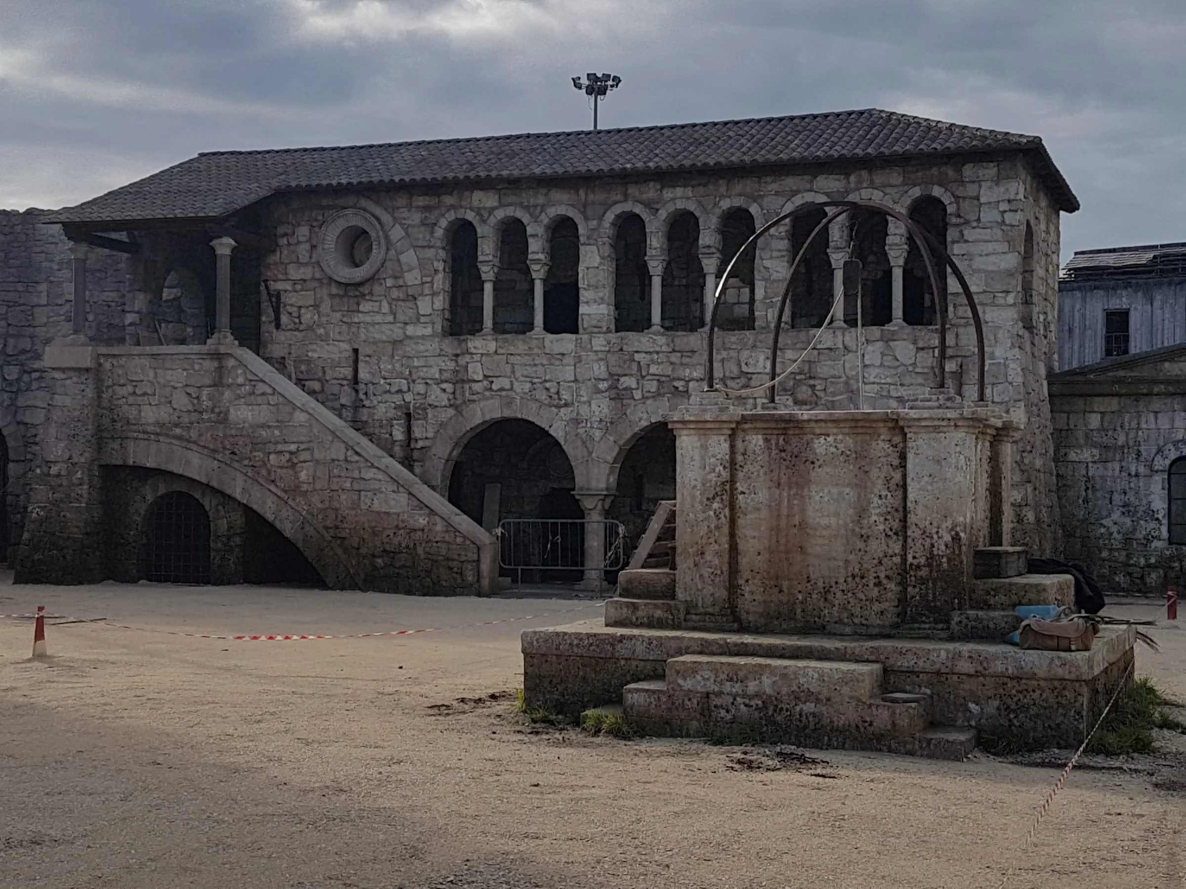 Il palazzo medievale italiano costruito da Creation Art per 6 Underground: edificio a due piani in finta pietra con bifore e trifore al piano superiore, scalinata esterna laterale, porticato a tre arcate, fontana ottagonale con balaustra al centro del cortile, terreno sabbioso del piazzale, cielo nuvoloso
