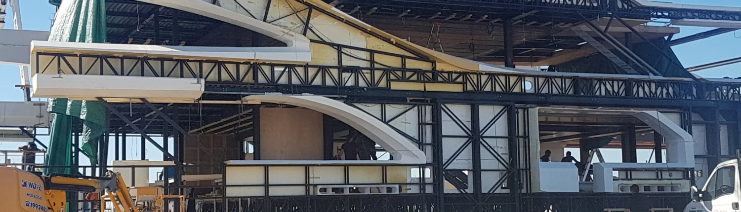 Vista panoramica dello scafo del mega-yacht visto di taglio: lunga struttura bianca a tre livelli con murata curva, struttura metallica nera a graticcio sui ponti, telone verde protettivo a sinistra, gru gialla in basso a sinistra e camion bianco a destra, sullo sfondo capannoni industriali