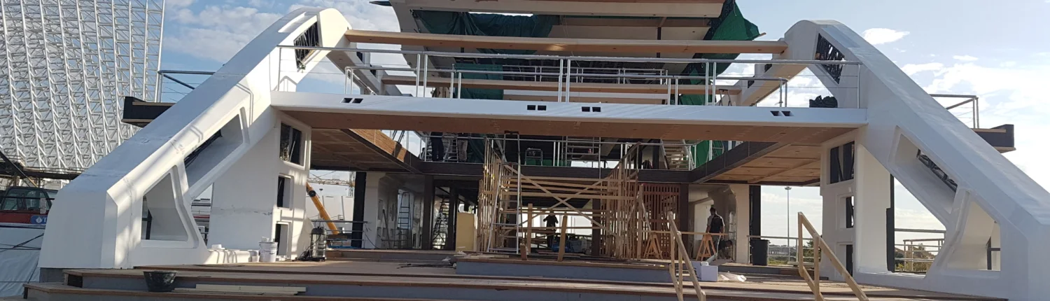 Altra vista panoramica dello yacht in piazzale: stessa struttura a tre livelli ma da angolazione diversa, profilo della murata che si snoda lungo tutta la lunghezza, telone protettivo, piazzale industriale