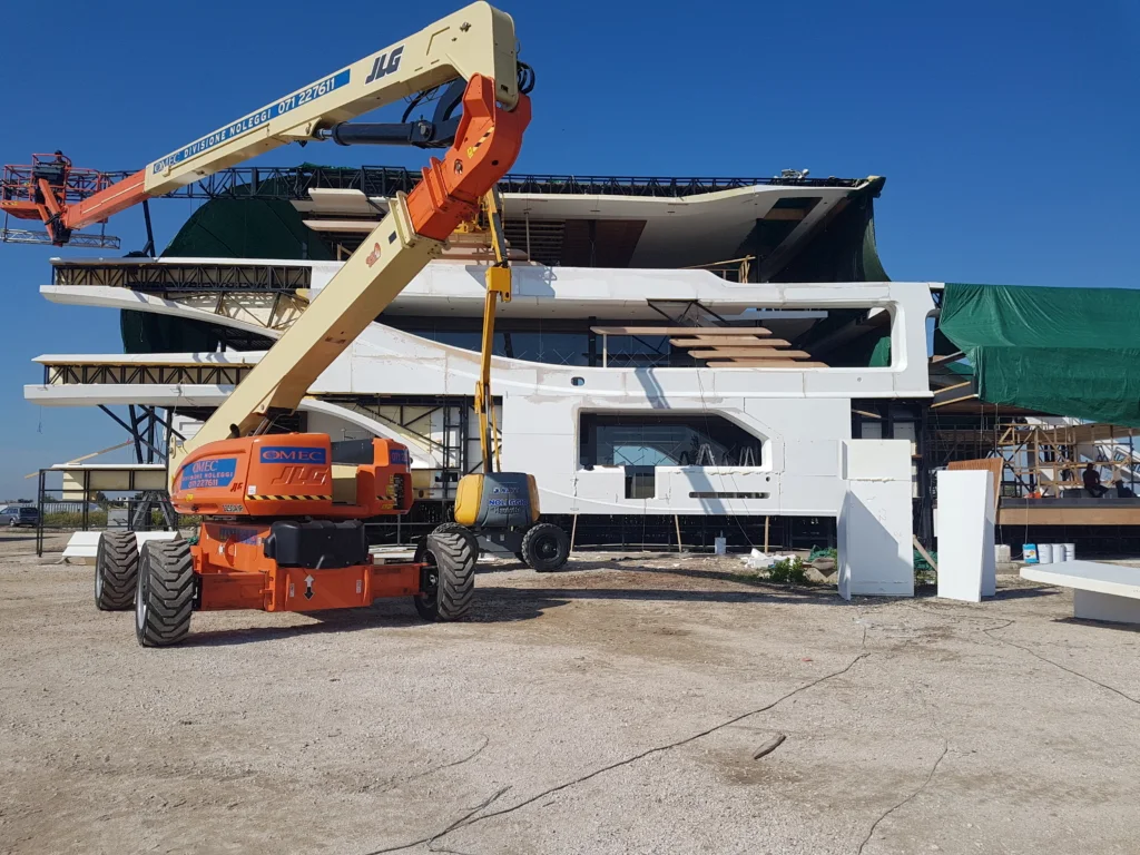 Il mega-yacht in fase di costruzione visto frontalmente nel piazzale: scafo bianco a tre piani con murata e ponti aperti, struttura interna in legno e graticcio metallico visibile, telone protettivo verde sui bordi, in primo piano una gru a piattaforma JLG arancione-bianca posizionata per le finiture in quota, cielo azzurro sgombro