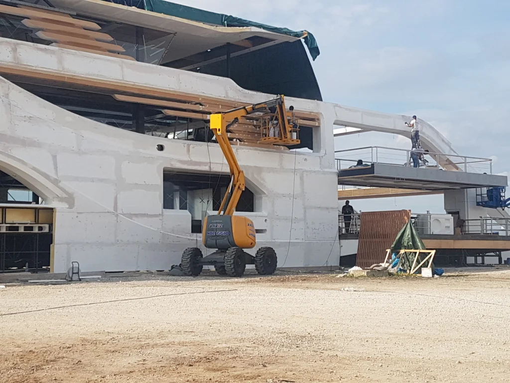 Dettaglio del piazzale di lavorazione: parte dello scafo bianco visibile in alto, in primo piano materiali del cantiere e pannelli di polistirolo bianchi appoggiati a terra, attrezzatura di lavorazione sparsa sul piazzale