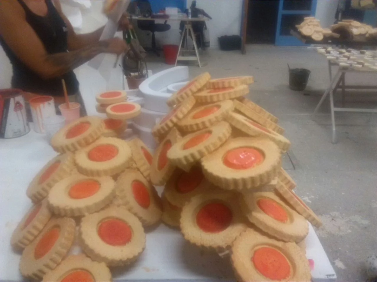 Giant cookies sculpted in polystyrene and fiberglass in the Creation Art workshop for the Lavazza commercial: stacks of round cookies with scalloped shortcrust edges, central orange/red circle simulating jam, a set designer painting them by hand in the background