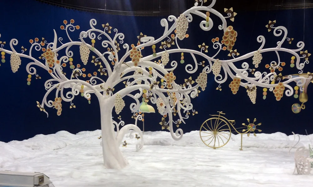The Lavazza set: large white liberty tree sculpted into sinuous curves with clusters of coloured coffee beans hanging from the branches like grapes, under an intense night-blue cyclorama, white snowy floor; in the foreground a gilded vintage penny-farthing, parasols and coloured pinwheels
