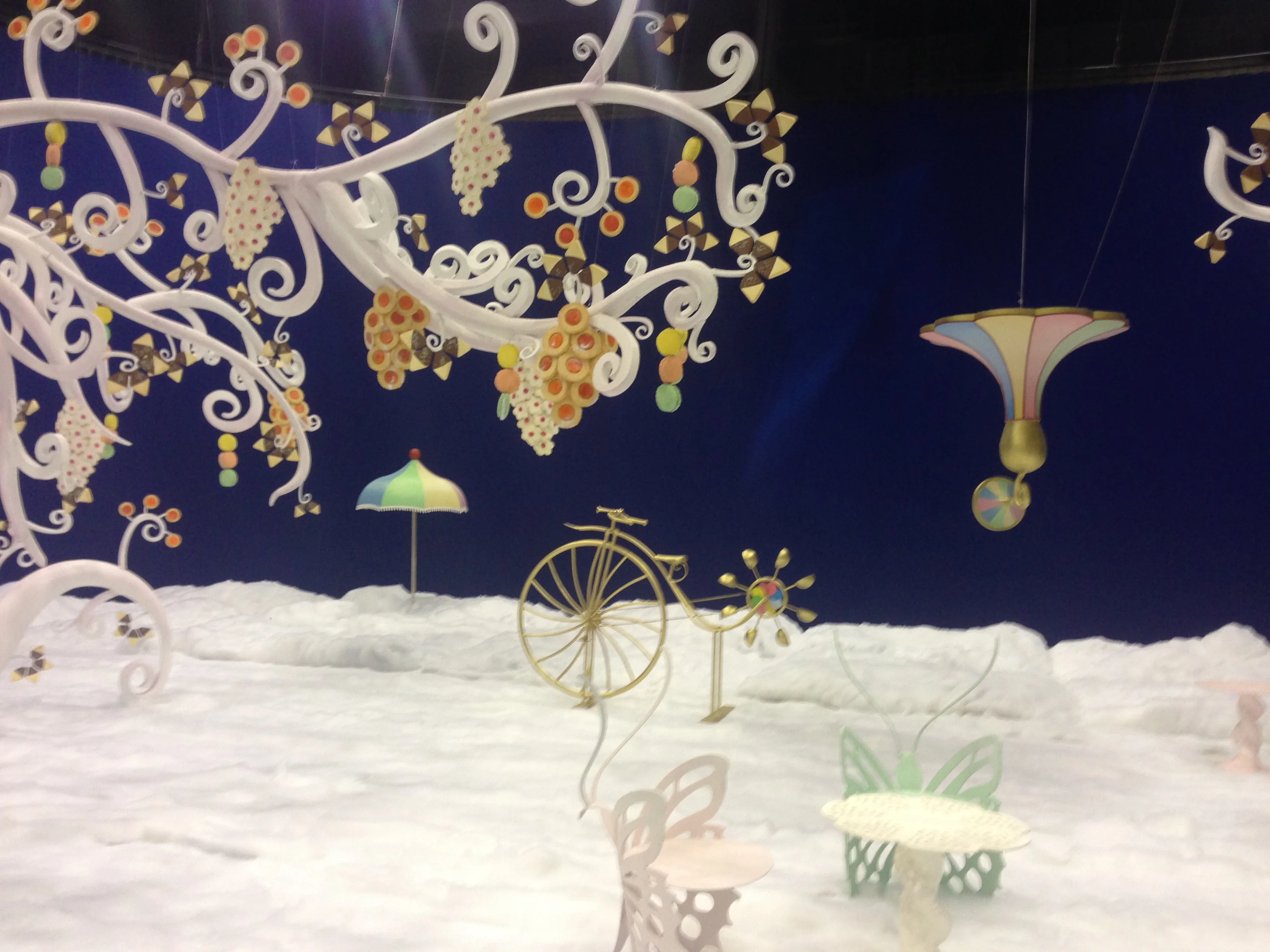 Lavazza set: stylised white tree with liberty curves and clusters of coloured coffee beans (green, orange, yellow) hanging like grapes; vintage yellow penny-farthing in the foreground; multicolour folding parasol; night-blue cyclorama backdrop, white snow on the ground, green and pink butterfly benches