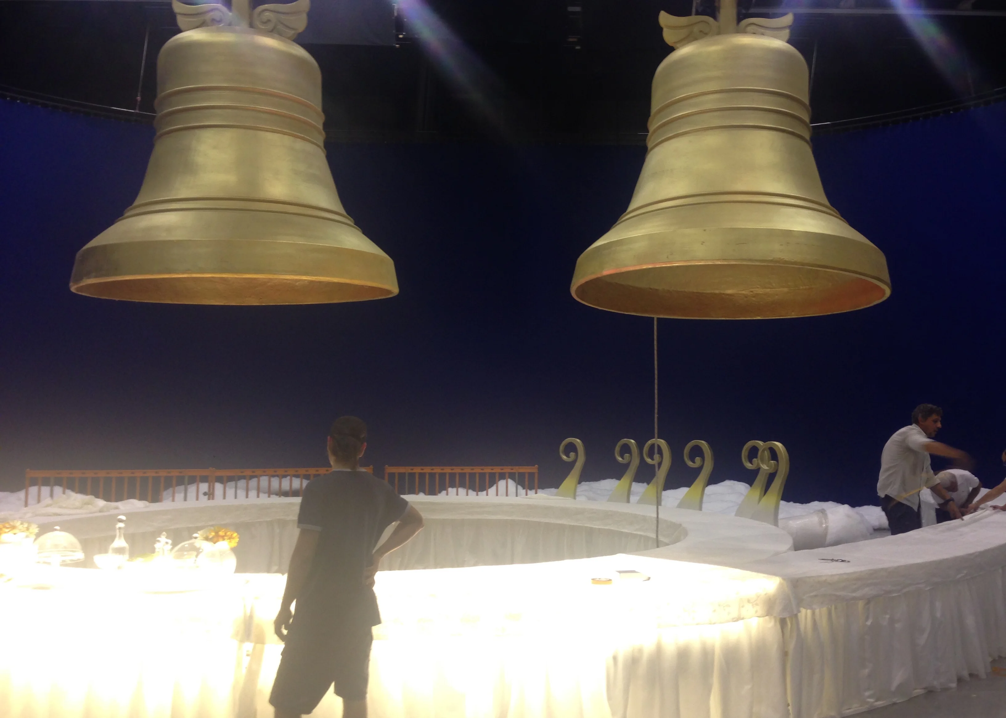 Two large golden bells suspended above a white inverted-C-shaped table on the Lavazza set; bells sculpted with small side wings, night-blue cyclorama in the background; a technician at work in the foreground in front of the table, other crew on the right