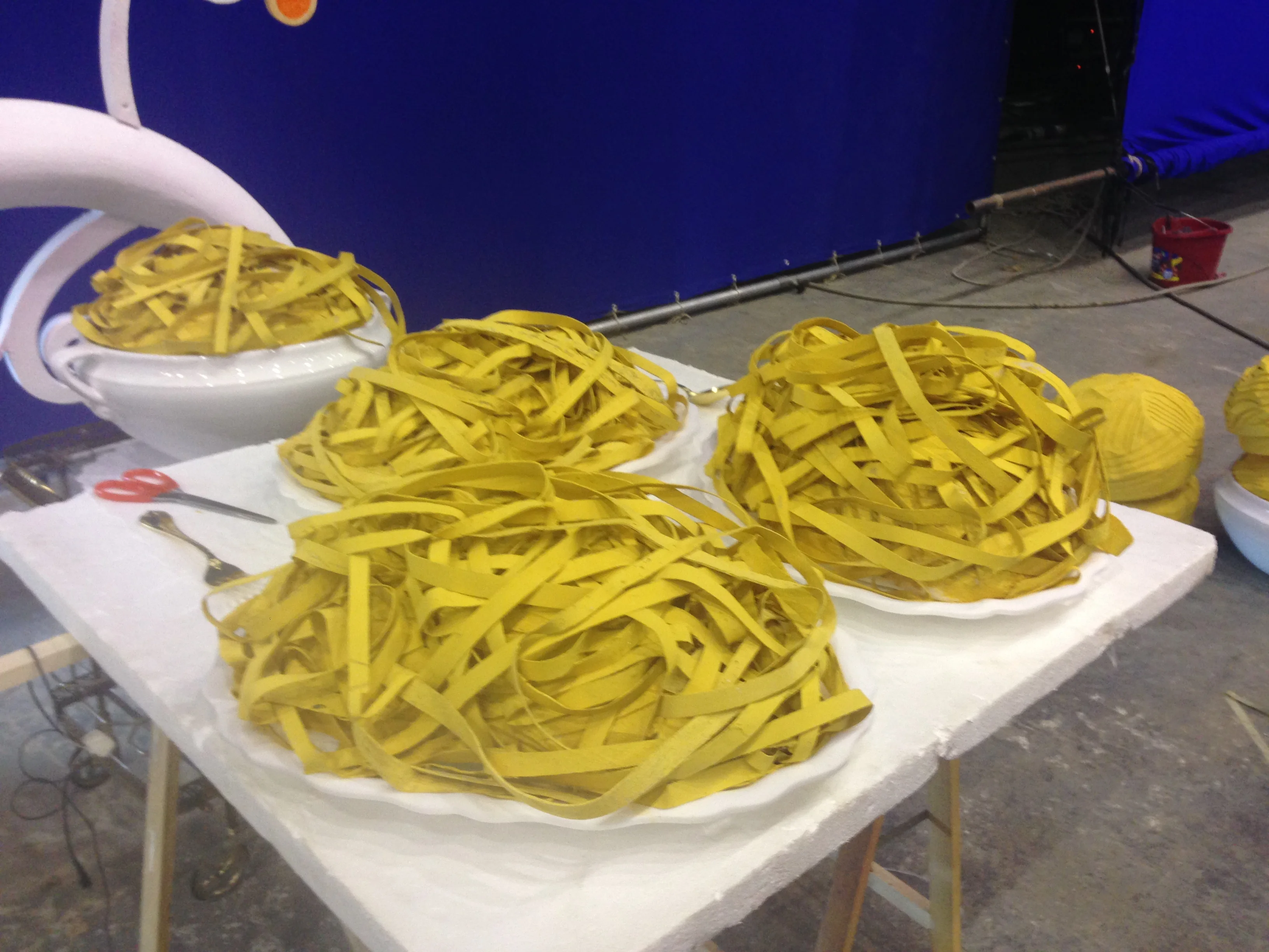 Giant tagliatelle plates sculpted for the Lavazza commercial: four large white plates topped with oversize yellow tagliatelle nests, each ribbon hand-modelled; blue cyclorama backdrop in the background