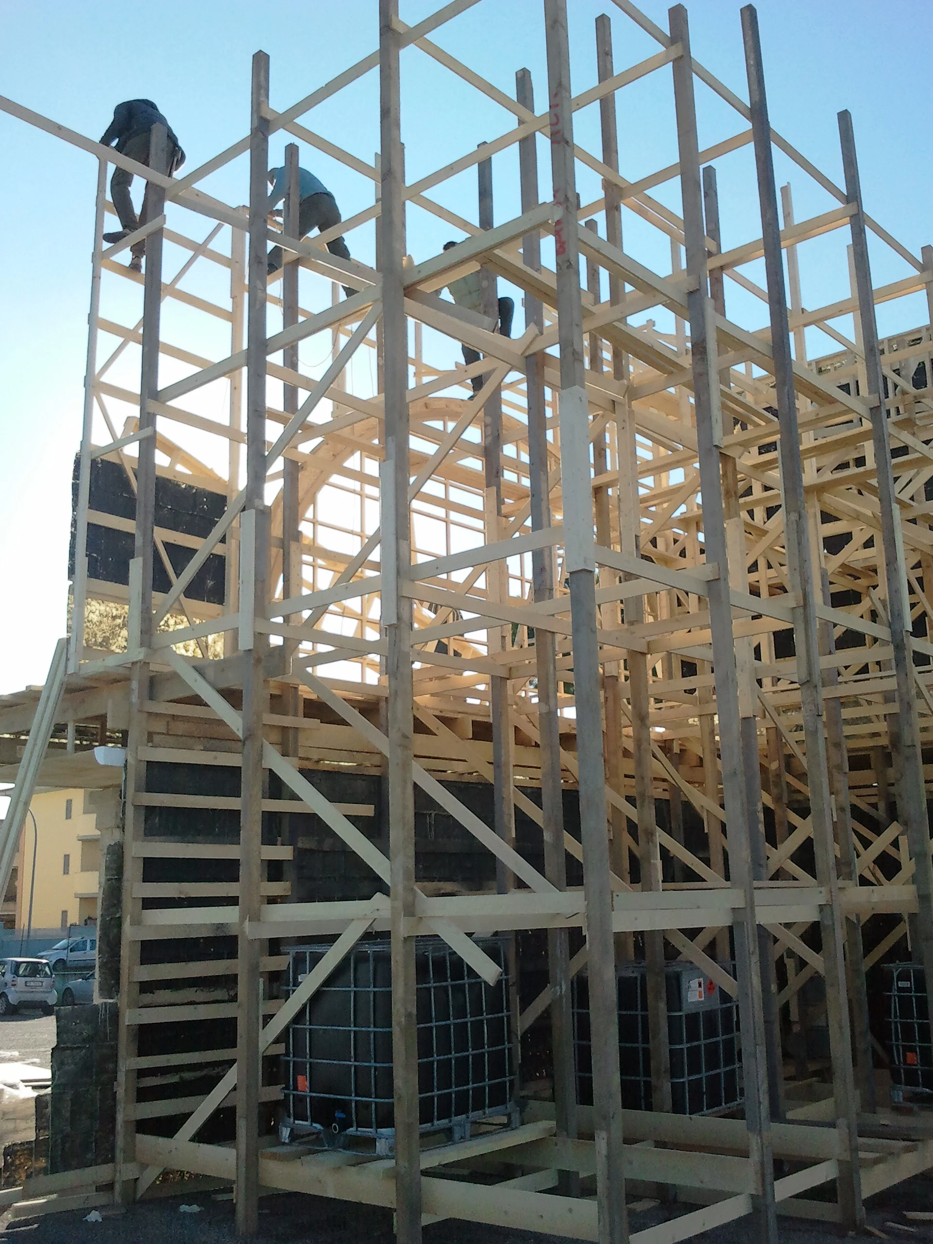 Construction of the Peroni set on 6 December 2012: three workers on the high scaffolds of the wooden structure; poles, horizontal scaffolding, crossed ladders, white containers at the base, blue sky