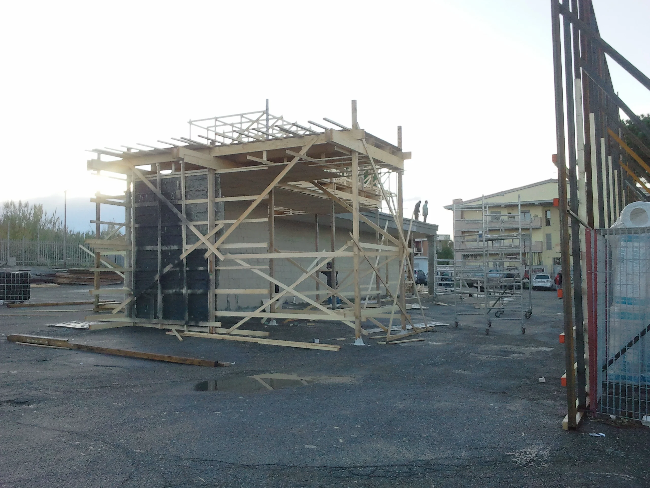 Construction of the Peroni set at dawn on 5 December 2012: skeleton wooden structure with the scaffold still empty, industrial buildings in the background, pink-blue dawn sky, paved yard