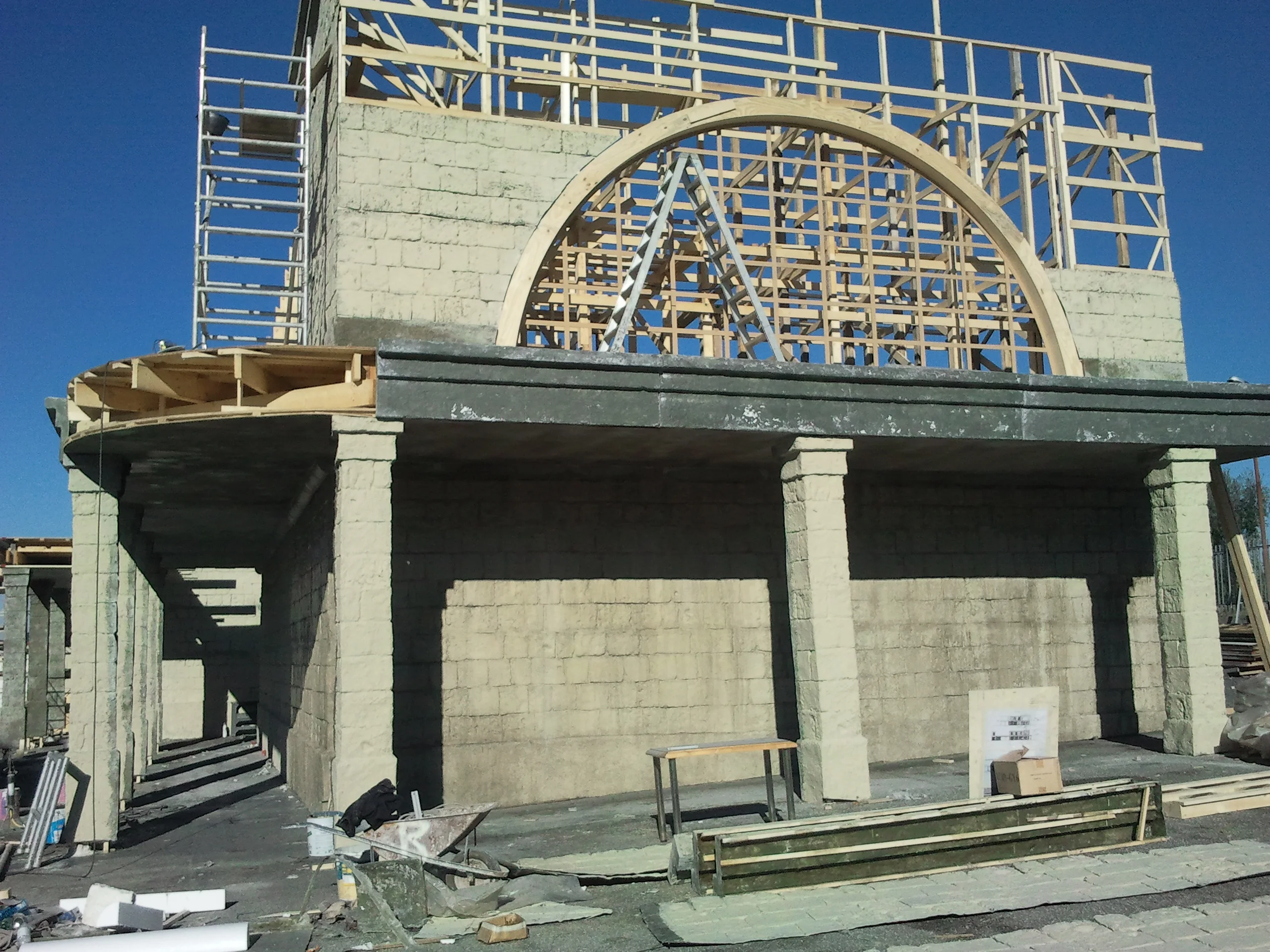 Side view of the Peroni construction: complete wooden structure with scaffolding, residential buildings in the background, parking lot with cars in front