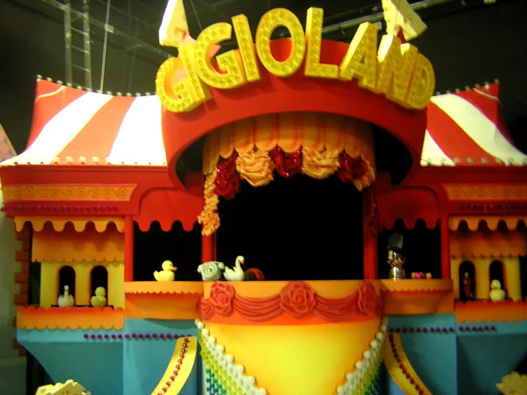 'Gigioland' set of the Topo Gigio Show: large red tent with white stripes in the centre, illuminated three-dimensional yellow 'GIGIOLAND' sign above it, central balcony with red drapes in garlands, white little animals (duck, swan, small knight) on the side niches, multicoloured rainbow staircase at the bottom, railings of beads and pink hearts