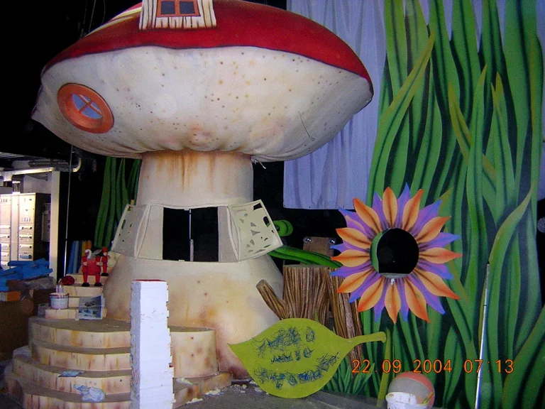 Topo Gigio Show set, 2004 season: large giant mushroom with red polka-dot cap, oval window and chimney like a house, beside it an oversize daisy-flower with orange and fuchsia petals, green dewdrops and a giant leaf on the ground, cartoon foliage painted on the studio walls, dated 22 September 2004