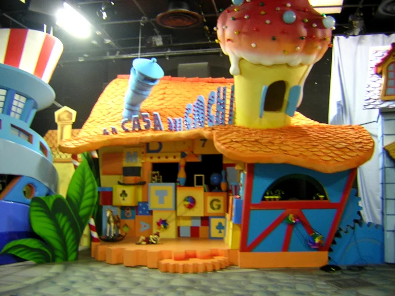 Side view of the same set: giant mushroom with red polka-dot cap, another smaller mushroom behind it, tall cartoon green grasses on either side, painted landscape in the background, cartoon carpet floor